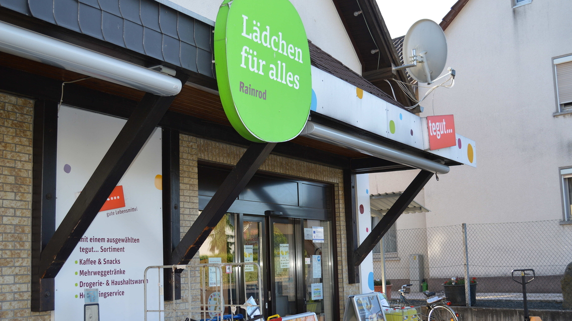 Die Fassade eines Tegut ladens mit einem grünen Schild, auf dem „Lädchen für alles“ steht, sowie der Standort Rainrod.