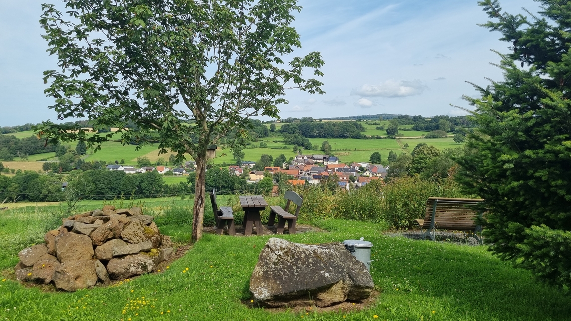 Blick auf eine grüne Wiese mit einem Baum, mehreren Bänken und einem großen Stein im Vordergrund und das Dorf Kaulstoß im Hintergrund.
