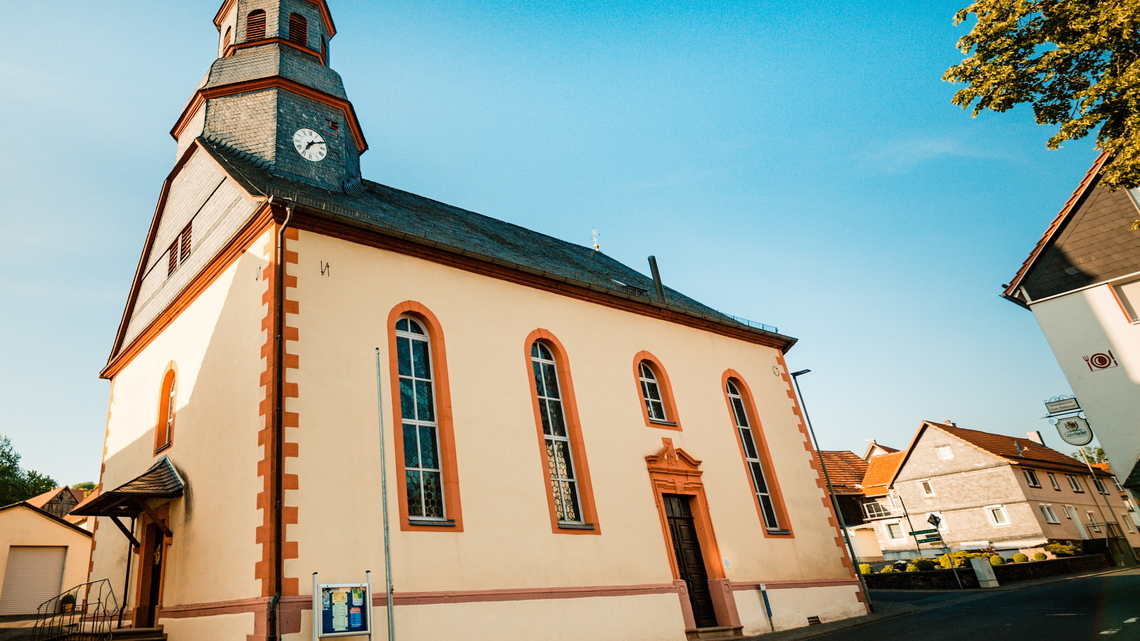 Dorfkirche in Burkhards mit Turmuhr, spitzbogigen Fenstern und hellem Putz mit roten Ecksteinen unter blauem Himmel.