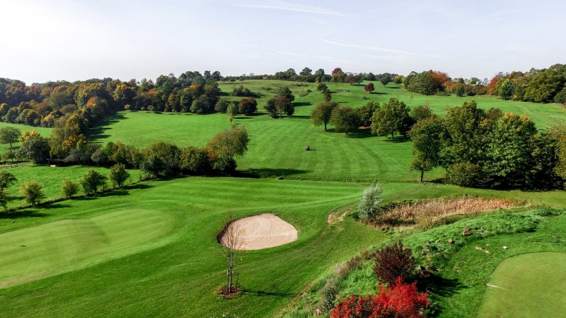 Grüne Golfplatzlandschaft mit gepflegtem Fairway, Sandbunker und Baumgruppen auf leicht hügeligem Gelände.