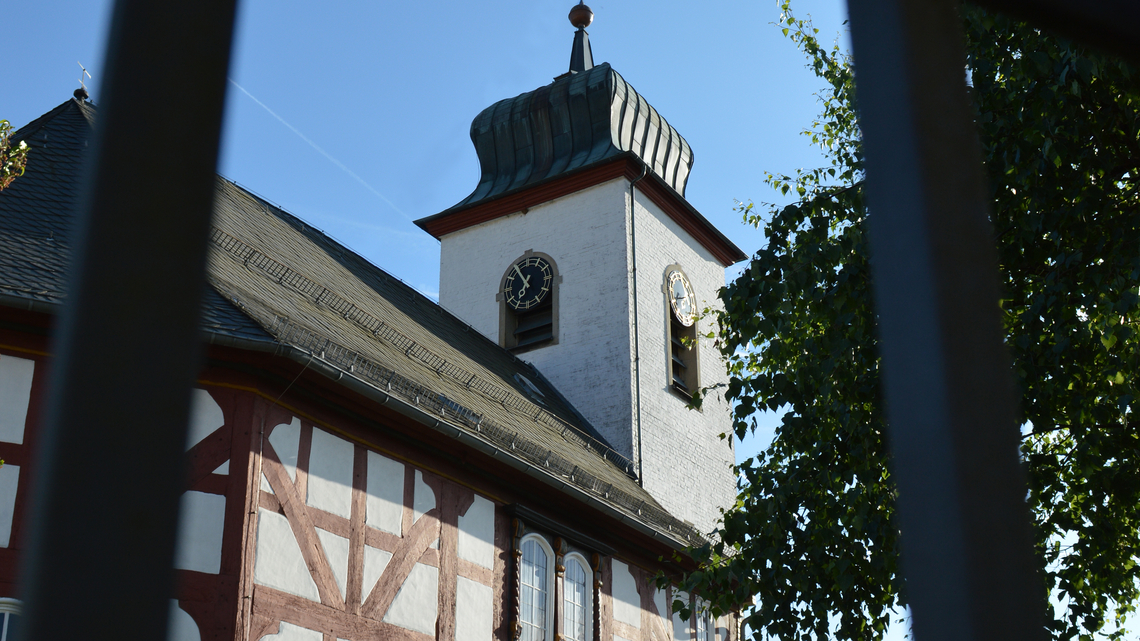 Kirchturm der Kirche in Breungeshain mit Zifferblättern an zwei Seiten und einem geschwungenen Dach, davor ein Fachwerkgebäude und ein Baum.