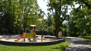 Spielplatz mit Klettergerüst, Rutsche und Sandfläche im Alteburgpark in Schotten, umgeben von Bäumen und einem gepflasterten Weg.