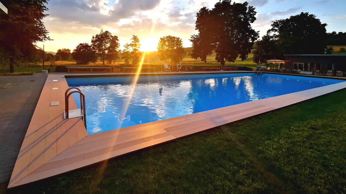 Freibad mit rechteckigem Schwimmbecken, umgeben von Rasenflächen und Bäumen bei Sonnenuntergang.
