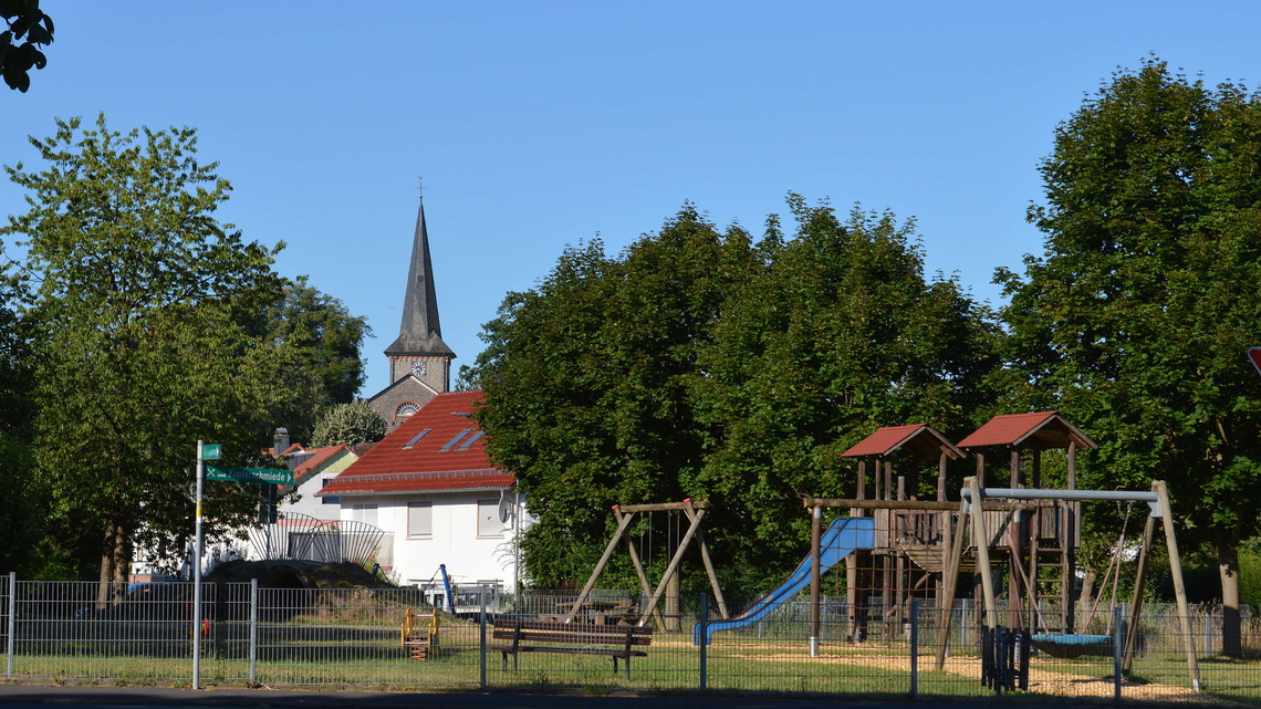 Ein Spielplatz mit Schaukeln, Rutsche und Klettergerüst unter strahlend blauem Himmel, umgeben von Bäumen und mit dem Kirchturm von Rainrod im Hintergrund.