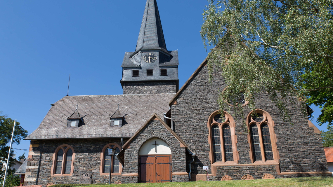 Eine historische Steinkirche in Wingerhausen mit einem hohen, grauen Kirchturm unter strahlend blauem Himmel.