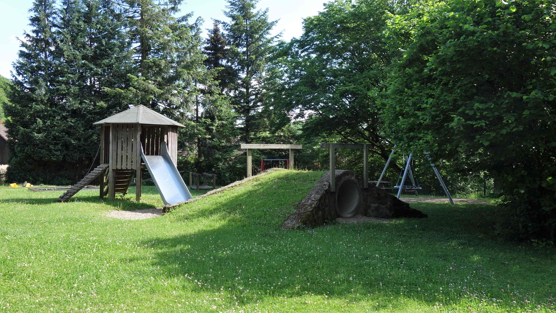 Kinderspielplatz mit Holzturm, Rutsche, Schaukeln und Kletterelementen auf einer Wiese umgeben von Bäumen.