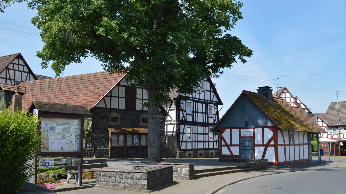 Fachwerk-Backhaus mit rotem Dach und Moosbewuchs neben einem großen Baum und einem Informationsschild in Eichelsachsen.