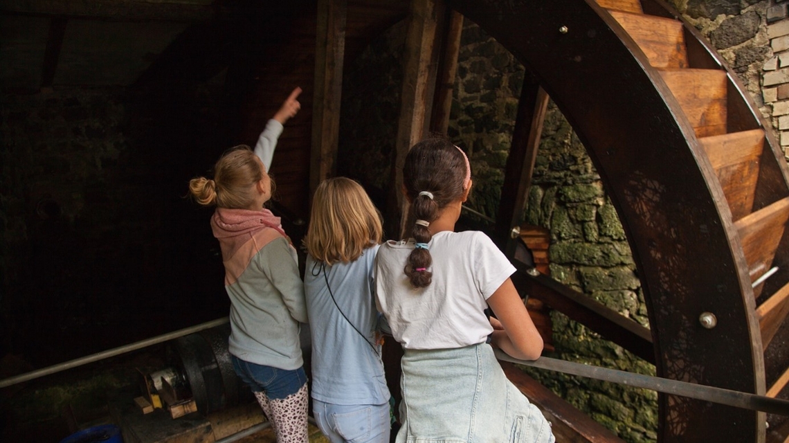Drei Kinder stehen vor einem großen hölzernen Wasserrad in der Weidmühle und blicken nach oben, eines zeigt mit dem Arm nach oben.