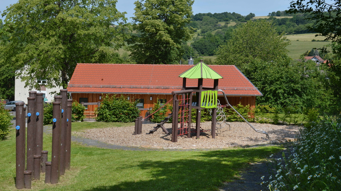 Kinderspielplatz mit Klettergerüst, Rutsche und Balancierbalken vor einem roten Gebäude und umgeben von Bäumen und grünen Hügeln.