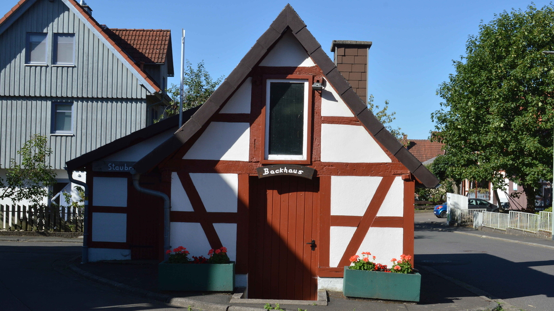 Fachwerkhaus mit roter Tür und Schild 'Backhaus' in Götzen, umgeben von weiteren Häusern und Pflanzen in Blumenkästen.