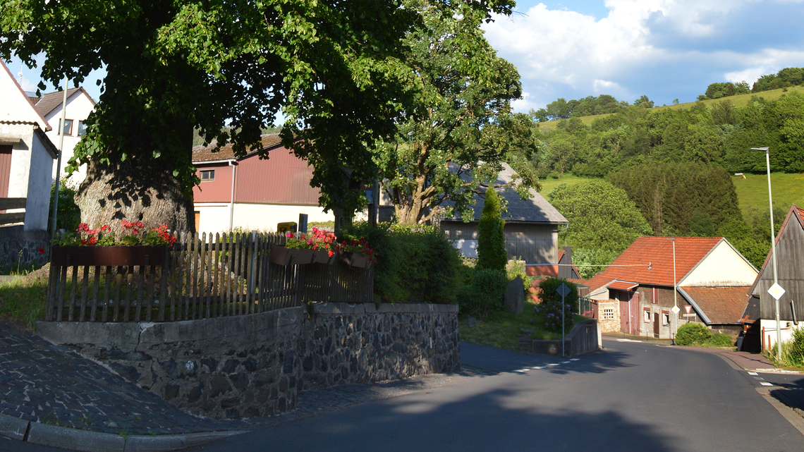 Eine friedliche Dorfszene mit malerischen Häusern, einen großen Baum, roten Blüten in Pflanzkübeln, üppigem Grün und einem strahlend blauen Himmel.