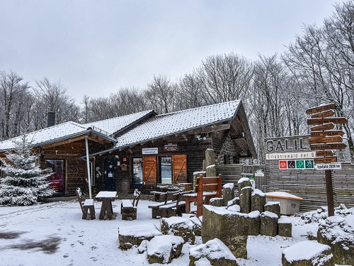 Schneebedecktes Holzhaus mit Außensitzbereich und Wegweiser in einem winterlichen Wald.