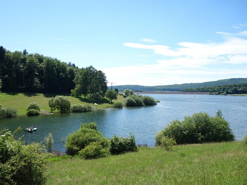 See mit Uferböschung, Büschen und Bäumen vor bewaldeten Hügeln unter blauem Himmel.