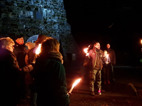 Gruppe von Menschen vor einer alten Steinmauer, die Fackeln in der Hand halten, bei Nacht.