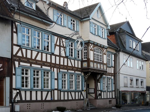Fachwerkhaus mit blauen Fensterläden und einem runden Uhrenschild an der Fassade in einer Straße mit weiteren Häusern.
