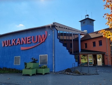 Das Vulkaneum in Schotten mit blauem Hauptgebäude, roter Aufschrift 'VULKANEUM' und angrenzendem orangefarbenem Gebäudeteil mit Turm.
