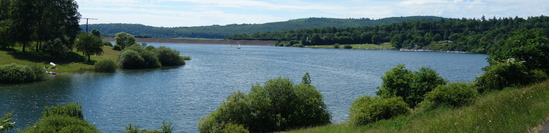See mit Uferböschung, Büschen und Bäumen vor bewaldeten Hügeln unter blauem Himmel.