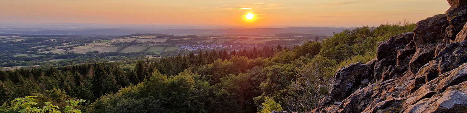 Sonnenuntergang über einer bewaldeten Landschaft mit Felsen im Vordergrund.